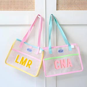 May include: Two mesh tote bags with pastel accents. One bag has yellow trim and the letters "LMR" in yellow. The other bag has green trim and the letters "CNA" in pink. Both bags have pink and blue handles.