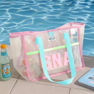 May include: A pink and white mesh beach bag with light blue handles and the word "CNA" in pink letters. The bag has a pink trim and a light green horizontal stripe. A bottle of lemonade sits to the left of the bag.