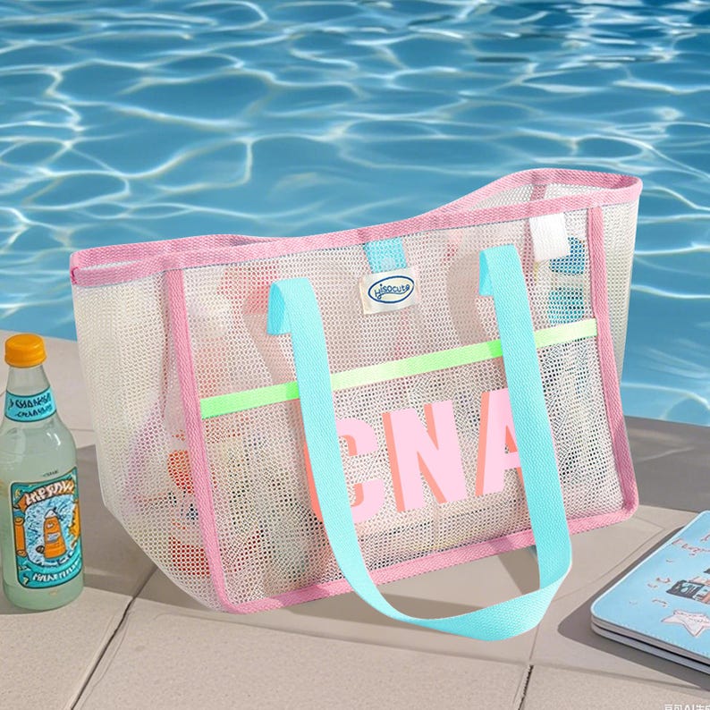 May include: A pink and white mesh beach bag with light blue handles and the word "CNA" in pink letters. The bag has a pink trim and a light green horizontal stripe. A bottle of lemonade sits to the left of the bag.