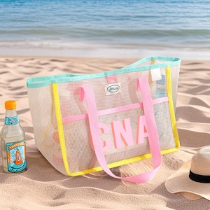 May include: A mesh beach tote bag with pink straps and the letters "CNA" in pink. The bag has a yellow and teal trim. A bottle of beverage and a straw hat are on the sand next to the bag.