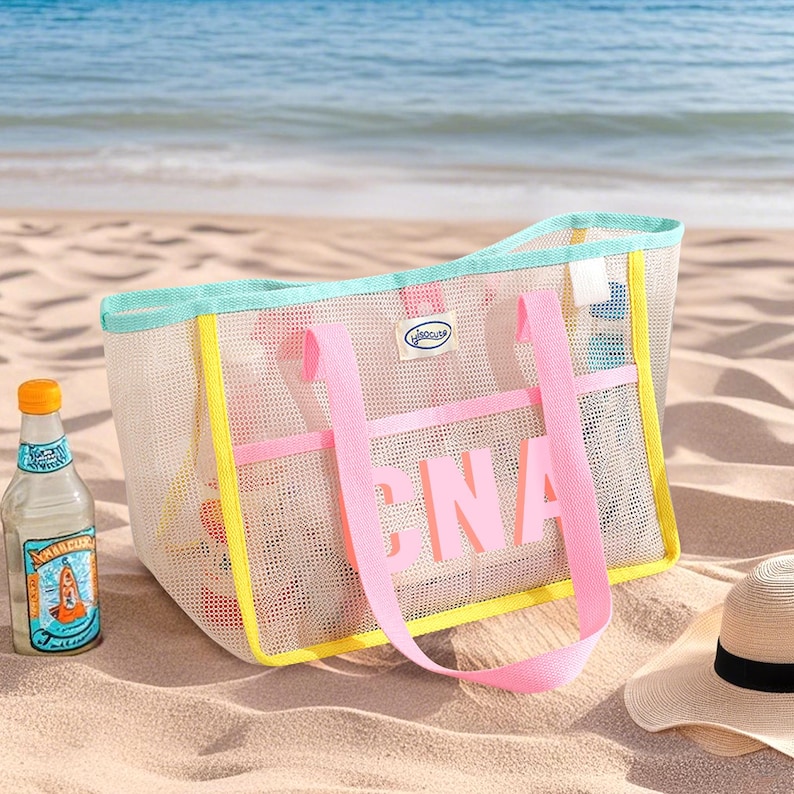 May include: A mesh beach tote bag with pink straps and the letters "CNA" in pink. The bag has a yellow and teal trim. A bottle of beverage and a straw hat are on the sand next to the bag.