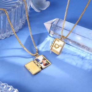 May include: Two gold-colored locket necklaces with rectangular pendants on a blue surface. One locket is open, revealing a photograph. The other locket has a heart and arrow design with the text "143" and "EL CORAZON YOU".