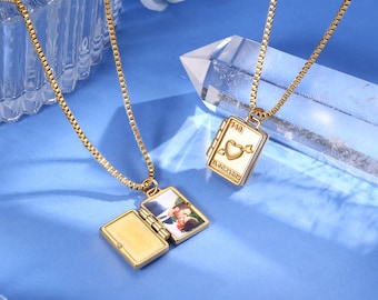 Puede incluir: Dos collares dorados con colgantes rectangulares en una superficie azul. Un relicario está abierto, revelando una fotografía. El otro tiene un diseño de corazón y flecha con el texto "143" y "EL CORAZON YOU".