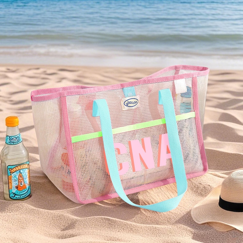 May include: A pink mesh beach tote bag with the word "PINA" in pink letters. The bag has light blue handles and a neon green stripe. A bottle of beverage and a straw hat are on the sand.