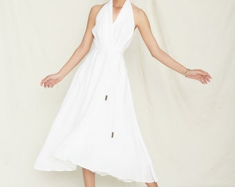 May include: A white halter dress with a flowing skirt. The dress features a wrap-around waist and is adorned with wooden beads on the tie. The model is wearing white sandals. The dress is made of a lightweight fabric.