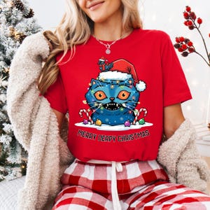 May include: Red t-shirt with a blue cat graphic wearing a Santa hat and Christmas lights. The cat has orange eyes and candy cane accents. The text "MERRY DERPY CHRISTMAS" is below the graphic.
