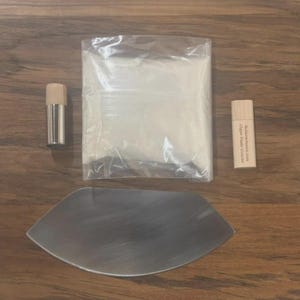 May include: An assortment of items including a stainless steel cheese knife, a small cylindrical container with a wooden top, a clear plastic bag, and a small wooden rectangular container with text. All items are on a wooden surface.