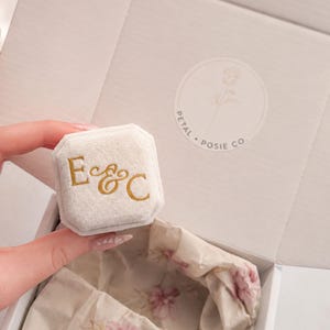 May include: An octagonal cream velvet ring box featuring the gold embossed letters E & C on the lid. The box is presented inside an open white gift box with a Petal Posie Co. logo and floral tissue paper lining the interior of the packaging.