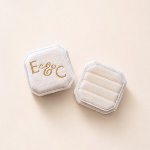May include: Two octagonal ring boxes covered in cream-colored velvet. One box lid features the gold-colored letters E & C in a serif font. The second box is open, revealing a cream-colored velvet interior with three horizontal slots for rings.