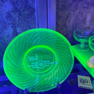 May include: A round, translucent green glass plate featuring a swirling radial pattern and the Vault-Tec logo etched in the center. The glass glows intensely under ultraviolet light, highlighting the circular design and the prominent brand text.