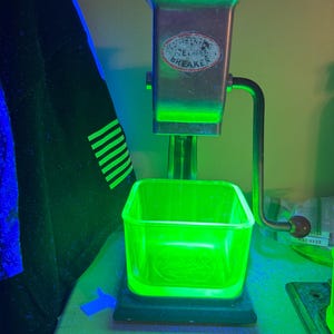 Puede incluir: Un triturador de hielo de metal vintage con manivela y un recipiente de vidrio cuadrado que brilla en verde brillante bajo luz ultravioleta. La parte frontal de la carcasa de metal tiene una etiqueta ovalada con el texto ICE BREAKER en letras oscuras.