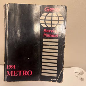 May include: A black 1991 Geo Metro service manual with pink text and a white globe graphic. The cover features the text Geo, Service Manual, 1991, METRO, and the code ST370-91. The book has a vertical column of gray rectangles on the right side.