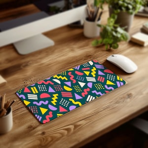 May include: A rectangular desk mat featuring a dark teal background with a repeating pattern of colorful geometric shapes including yellow triangles, pink squiggly lines, purple semicircles, and white dotted squares.