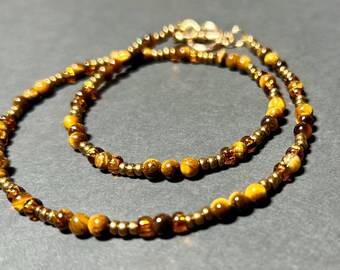 May include: A beaded necklace featuring tiger's eye and gold-colored beads. The necklace is composed of small, round beads in shades of brown and gold, interspersed with larger, polished tiger's eye beads. The necklace is secured with a gold-toned clasp.