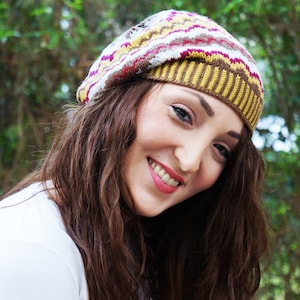 May include: A colorful knitted beret with a zig-zag pattern in shades of yellow, brown, pink, and white. The beret is worn by a person with long brown hair and a white shirt. The background is a blurred view of green foliage.