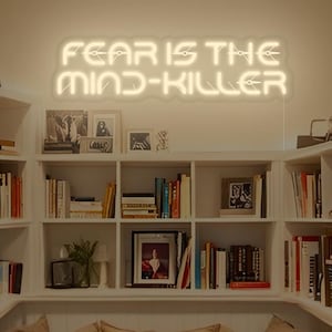 May include: A cozy reading nook with built-in bookshelves filled with books. Above, a neon sign reads "FEAR IS THE MIND-KILLER." The seating area features beige pillows and a neutral-toned mattress, creating a relaxing space for reading and contemplation.