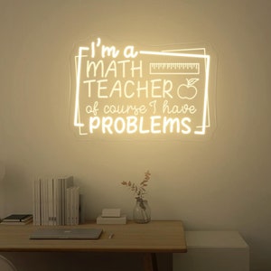 Op de afbeelding: Een wit neonbord met de tekst "Ik ben een wiskundeleraar, natuurlijk heb ik problemen" in een wit kader met een liniaal en een appelafbeelding.