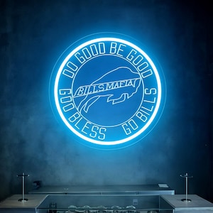 以下が含まれることがあります： 「DO GOOD BE GOOD」と「GO BILLS」の文字が書かれた青いネオンサイン。その下に「BILLS MAFIA」のロゴがあります。サインの下には、さまざまなTシャツが並んだディスプレイケースがあります。背景は濃い青色の壁です。