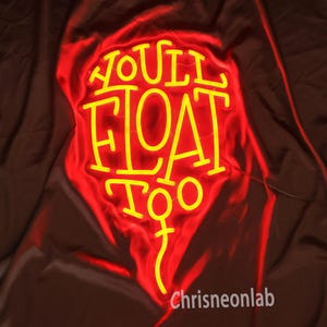Könnte beinhalten: Ein gelbes Neon-Schild mit dem Text "YOU'LL FLOAT TOO" vor rotem Hintergrund. Das Schild hat eine einzigartige Form und liegt auf einem dunkelbraunen Stoff. Der Text "Chrisneonlab" ist unten zu sehen.