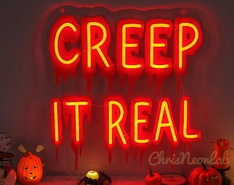 Op de afbeelding: Een rood neonbord met een gele omtrek waarop "Creep It Real" staat. Het bord hangt aan een muur met een witte achtergrond. Op de voorgrond staan kleine pompoenen en een spinnenbeeldje.