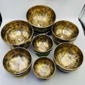 Puede incluir: Conjunto de ocho cuencos cantores, hechos de metal, con una variedad de tamaños y patrones. Los cuencos están dispuestos en un círculo sobre una superficie blanca. Cada cuenco tiene un diseño único y está decorado con intrincados patrones.