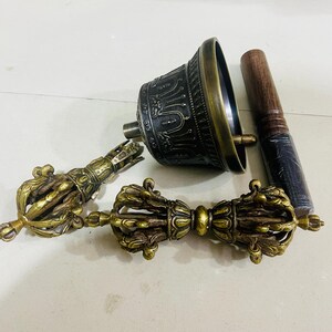 May include: A set of three Tibetan Buddhist ritual instruments: a hand bell, a dorje, and a wooden striker. The bell is made of brass and has intricate carvings. The dorje is a double thunderbolt, also made of brass. The striker is made of wood and has a dark brown finish.