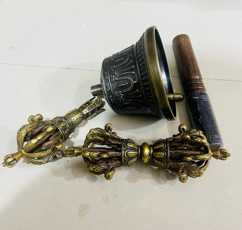 May include: A set of three Tibetan Buddhist ritual instruments: a hand bell, a dorje, and a wooden striker. The bell is made of brass and has intricate carvings. The dorje is a double thunderbolt, also made of brass. The striker is made of wood and has a dark brown finish.