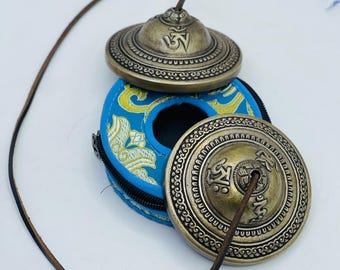 May include: A set of two small, brass-colored hand cymbals with a blue and gold embroidered pouch. The cymbals have a raised design and the word "Om" inscribed on them. The pouch has a zipper closure and a leather cord.