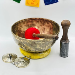 May include: A brown singing bowl with a yellow cushion, a red mallet, and two small brass cymbals. The bowl has a decorative design and the cymbals have Sanskrit writing.