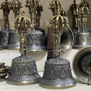May include: A collection of antique-looking, ornate, brass bells with intricate designs and a golden finish. The bells have a handle with a crown-like design and are decorated with small, raised figures.