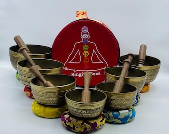 May include: A set of seven singing bowls with wooden strikers, each bowl resting on a colorful cushion. The bowls are arranged in a semi-circle around a red carrying case with a depiction of a meditating figure and the text "Singing Bowl".