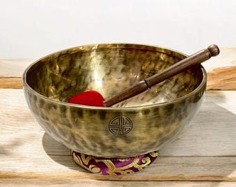 May include: A large, antique-looking brass singing bowl with a wooden striker and a red felt mallet. The bowl is resting on a purple and gold cushion with a traditional Chinese symbol on the side.