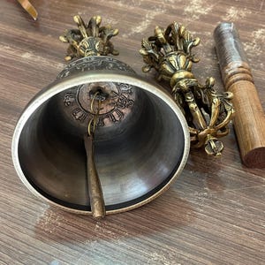 May include: A Tibetan singing bowl with a brass finish and intricate carvings. The bowl is accompanied by a wooden striker and a hand-held bell with a brass finish and intricate carvings.