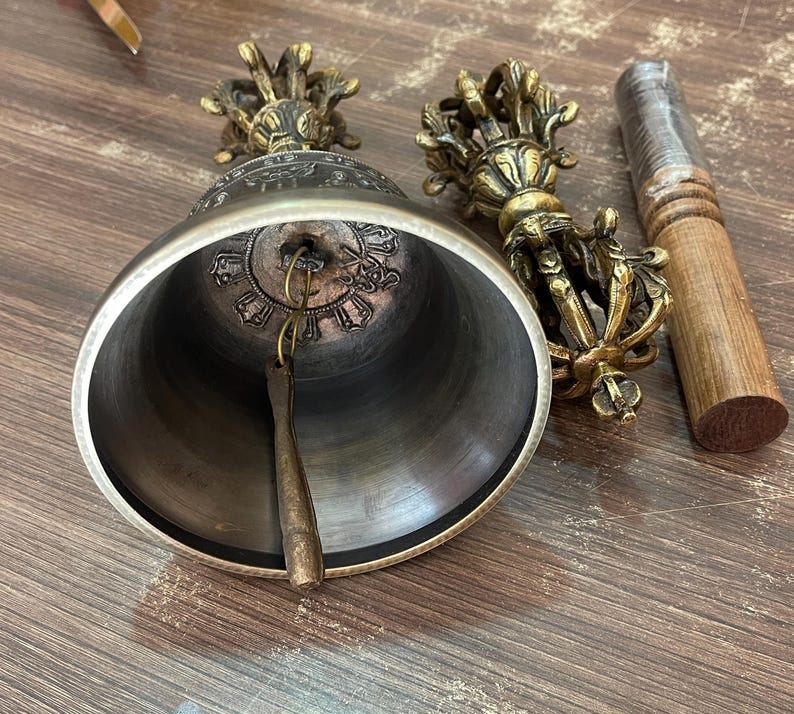 May include: A Tibetan singing bowl with a brass finish and intricate carvings. The bowl is accompanied by a wooden striker and a hand-held bell with a brass finish and intricate carvings.