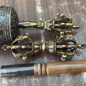 May include: Three ornate golden prayer wheels with intricate designs and a wooden prayer stick. The prayer wheels are used in Tibetan Buddhism to spin mantras and accumulate merit.