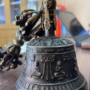 May include: A bronze Tibetan singing bowl with a detailed design and a small figure on top. The bowl has a handle with a decorative design.