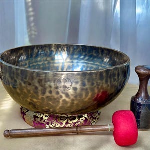 Pode incluir: Uma grande tigela de canto de cor bronze com um emblema decorativo, repousando sobre uma almofada estampada. Inclui-se um malho de madeira com uma cabeça de feltro vermelho e um batedor de madeira. A tigela é projetada para cura sonora e meditação.