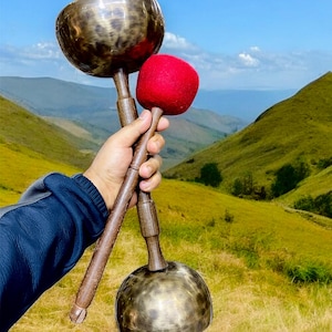May include: Two singing bowls, one larger than the other, with a wooden striker. The bowls are made of metal and have a dark brown colour. The striker is made of wood and has a red tip.