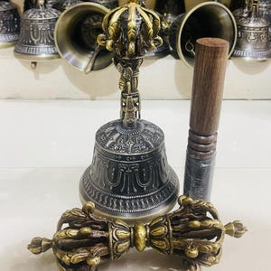 May include: A set of three Tibetan Buddhist ritual instruments: a hand bell with a decorative crown-shaped handle, a wooden striker, and a dorje, a ritual scepter with a double thunderbolt design. The bell and dorje are made of metal with intricate carvings and a dark patina.