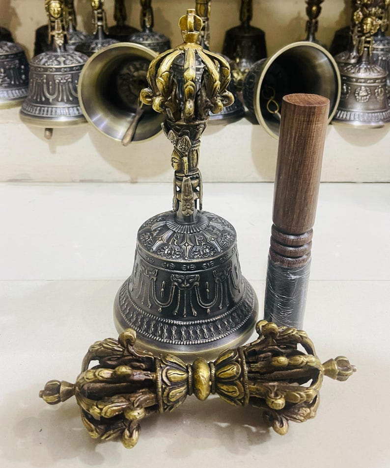 May include: A set of three Tibetan Buddhist ritual instruments: a hand bell with a decorative crown-shaped handle, a wooden striker, and a dorje, a ritual scepter with a double thunderbolt design. The bell and dorje are made of metal with intricate carvings and a dark patina.