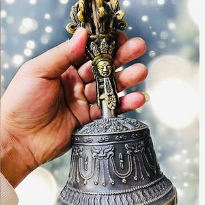 May include: A silver and gold bell with intricate carvings and a deity figure on the handle. The bell is held in a hand.