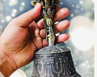 May include: A silver and gold bell with intricate carvings and a deity figure on the handle. The bell is held in a hand.