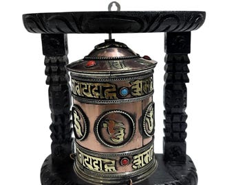 May include: A copper prayer wheel with gold lettering and colorful stones, mounted on a black wooden stand. The prayer wheel has the words "Om Mani Padme Hum" inscribed on it.