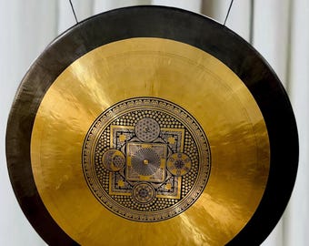 May include: A large, circular gong with a polished gold center and a dark brown rim. The center features an intricate black geometric design. The gong is suspended from a black metal bar by thin black cords.