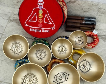 May include: A collection of brass singing bowls with various chakra symbols, a red cushion with a white illustration of a person and the text "Singing Bowl", and wooden strikers. The bowls are in different sizes.