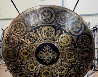 May include: A large, round gong with intricate gold-colored patterns on a black background. The design features various symbols and circular motifs, suspended from a black metal frame. The gong is likely used for sound healing or meditation.