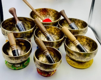 May include: Seven antique-looking singing bowls with wooden strikers. The bowls are made of metal and have a hammered finish. They are arranged in a semi-circle on a white surface. Each bowl has a unique design and inscription.