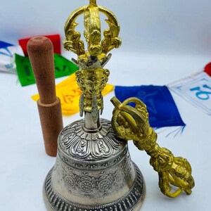 May include: A silver bell with ornate detailing, a gold-colored handle, and a wooden striker. The bell is accompanied by a gold-colored vajra. Colorful prayer flags are in the background. This item is likely used for spiritual or ceremonial purposes.