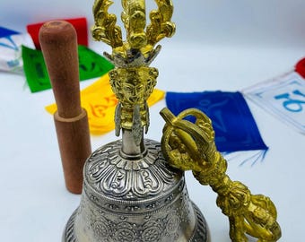 May include: A silver bell with ornate detailing, a gold-colored handle, and a wooden striker. The bell is accompanied by a gold-colored vajra. Colorful prayer flags are in the background. This item is likely used for spiritual or ceremonial purposes.