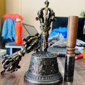 May include: A golden Tibetan singing bowl with intricate carvings and a matching hand-held striker. The bowl has a dark bronze finish and is decorated with a deity face and other symbolic figures. The striker is made of wood and has a black and brown finish.